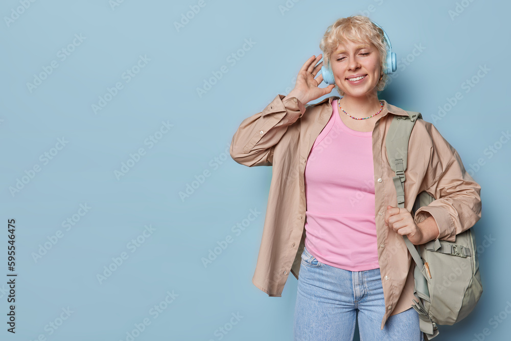 Horizontal shot of pleased blonde woman enjoys favorite playlist in headphones poses with rucksack on shoulder dressed casually ready for trip isolated over blue background copy space aside. Horizontal shot of pleased blonde woman enjoys favorite playlist in headphones poses with rucksack on shoulder dressed casually ready for trip isolated over blue background copy space aside.