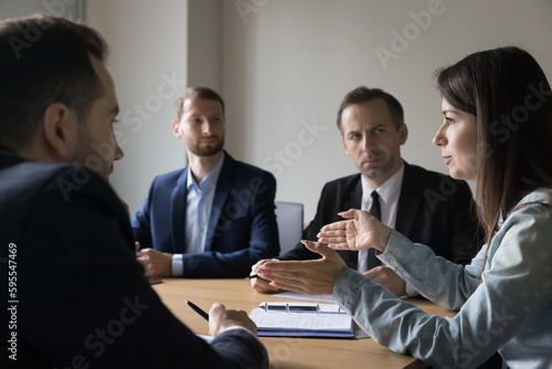 Confident female CEO speaking to coworkers on meeting, sharing ideas, negotiating on project, startup strategy. Young businesswoman sitting at conference table, talking to serious team