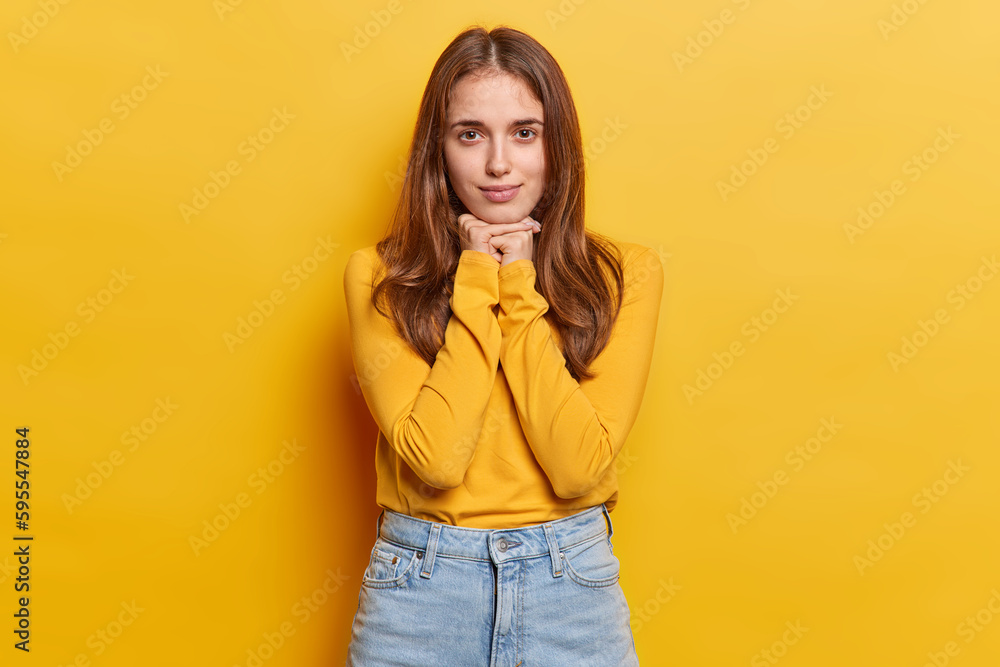 Studio shot of millennial girl keeps hands under chin has dark eyes long straight hair wears casual jumper and jeans focused directly at camera isolated on yellow background. Human facial expressions Studio shot of millennial girl keeps hands under chin has dark eyes long straight hair wears casual jumper and jeans focused directly at camera isolated on yellow background. Human facial expressions