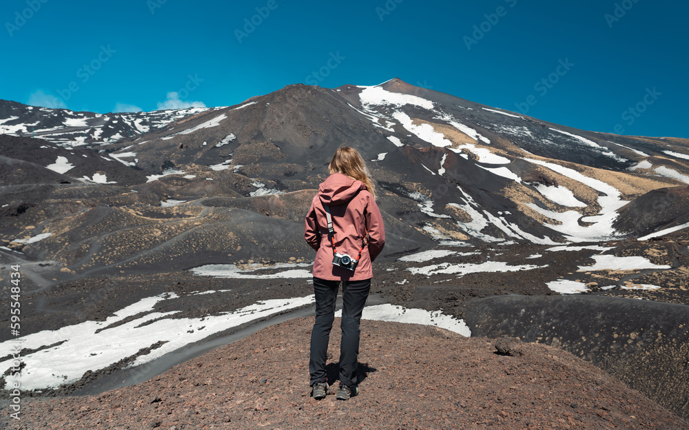Obraz premium Blond photographer observes the panorama of southern slope of the Etna volcano. Back view of unrecognizable hiker with shoulder camera getting inspired to take a landscape photo. Adventure and outdoor