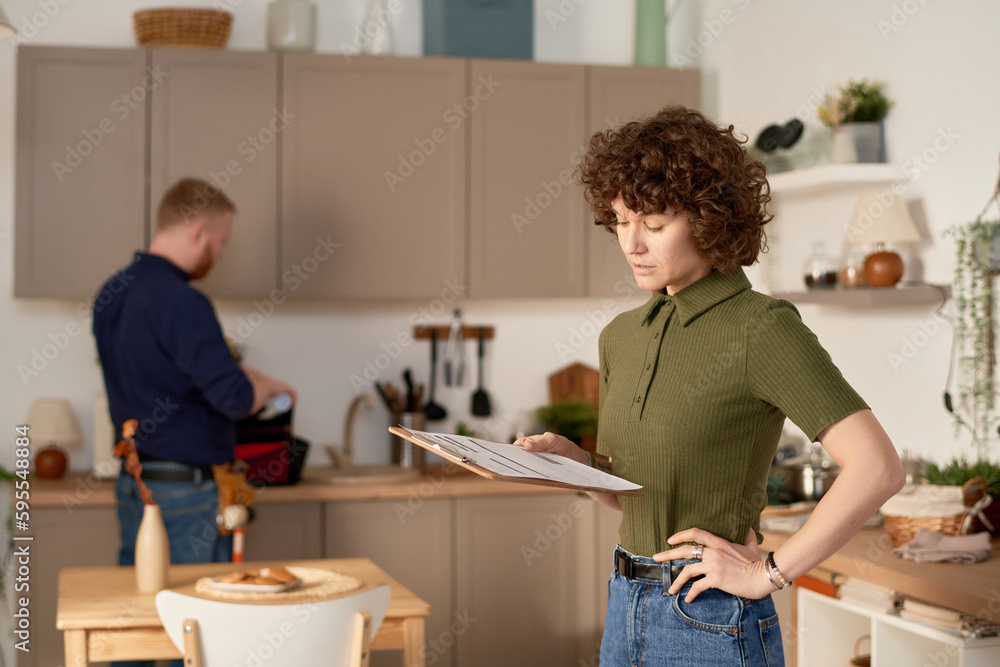 Housewife examining customer service contract standing in the kitchen with repairman working in the background Housewife examining customer service contract standing in the kitchen with repairman working in the background