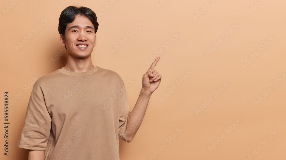 Horizontal shot of handsome cheerful Chinese man points finger right shows way demonstrates promo on copy space or product banner smiles toothily wears t shirt isolated over brown background Horizontal shot of handsome cheerful Chinese man points finger right shows way demonstrates promo on copy space or product banner smiles toothily wears t shirt isolated over brown background