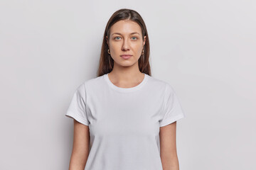 Waist up shot of nice looking woman has dark hair dressed in basic t shirt keeps arms down looks directly at camera isolated on white background. Brunette female model poses in studio for making photo