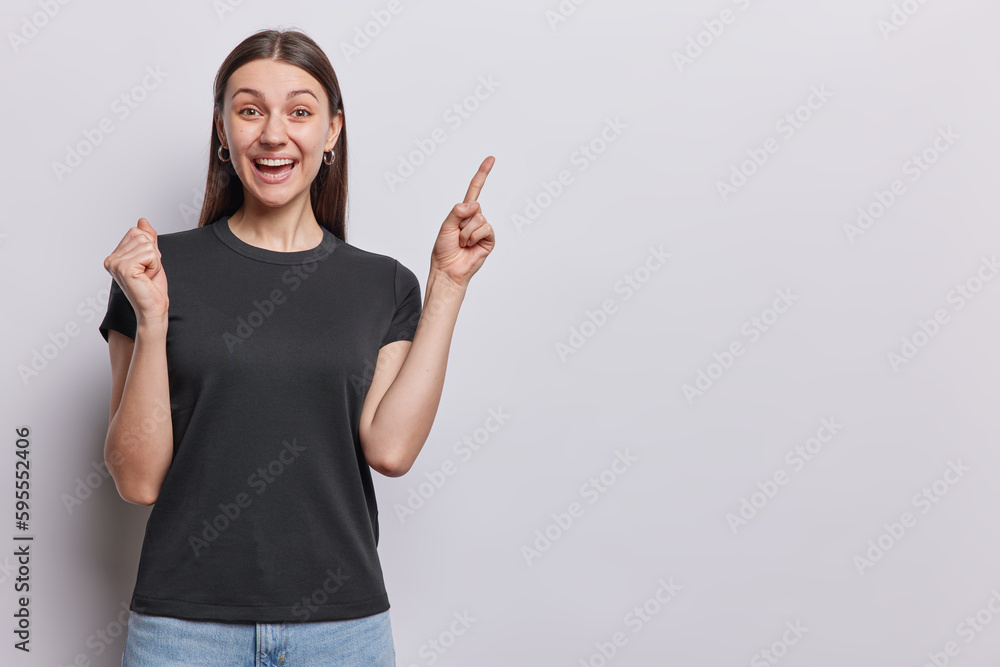 Horizontal shot of positive dark haired woman feels very happy clenches fist and points inndex finger at upper side dressed in casual black t shirt and jeans isolated over white wall. Hey look there Horizontal shot of positive dark haired woman feels very happy clenches fist and points inndex finger at upper side dressed in casual black t shirt and jeans isolated over white wall. Hey look there