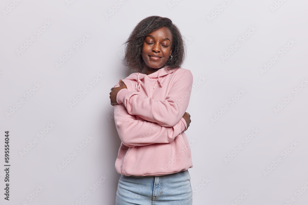 Good feelings and self love concept. Pleased curly haired Afro American girl embraces herself feels cozy in new comfortable sweatshirt stands with eyes closed isolated over white background. Good feelings and self love concept. Pleased curly haired Afro American girl embraces herself feels cozy in new comfortable sweatshirt stands with eyes closed isolated over white background.