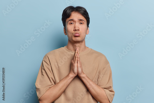 Obraz na plátně Intense handsome Asian man presses hands together in pray begs for something purses lips dressed in casual beige jumper isolated over blue background asks for forgiveness