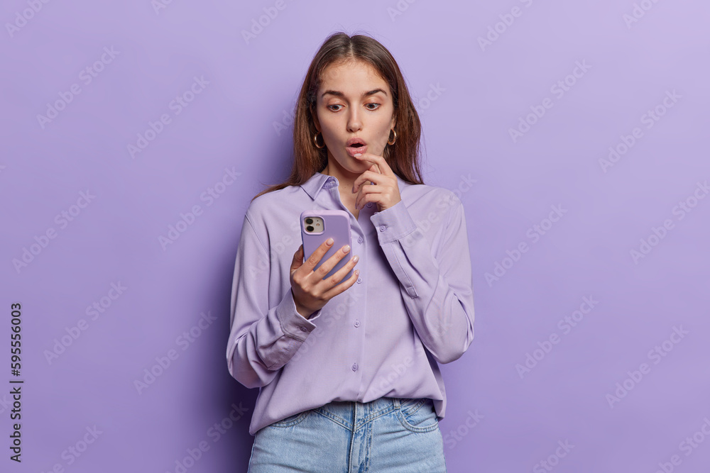 Studio shot of brunette woman keeps mouth opened has shocked expression stares in smartphone screen wears shirt and jeans finds out amazing news isolated over purple background. Omg concept. Studio shot of brunette woman keeps mouth opened has shocked expression stares in smartphone screen wears shirt and jeans finds out amazing news isolated over purple background. Omg concept.