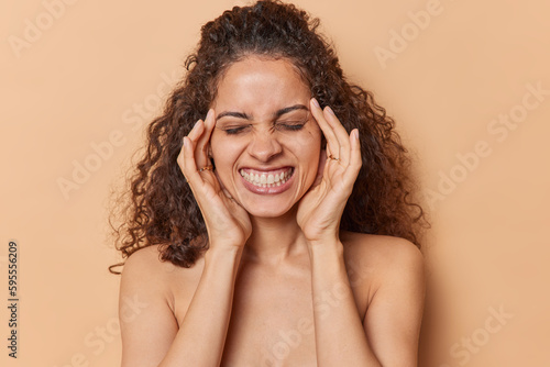 Headshot of dark haired young woman keeps hands on temples clenches teeth and closes eyes has healthy glowing skin stands bare shoulders isolated over brown studio background. Wellness concept