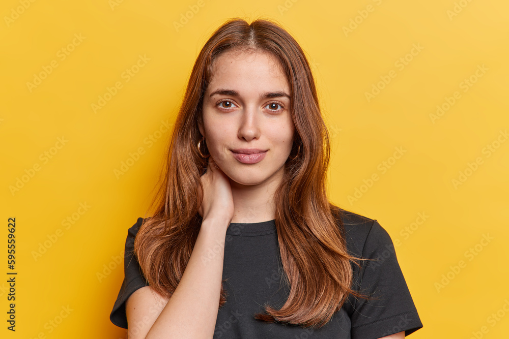 Portrait of good looking woman with dark straight hair dressed in casual black t shirt keeps hands on neck looks directly at camera isolated over vivid yellow background. People and face expression Portrait of good looking woman with dark straight hair dressed in casual black t shirt keeps hands on neck looks directly at camera isolated over vivid yellow background. People and face expression