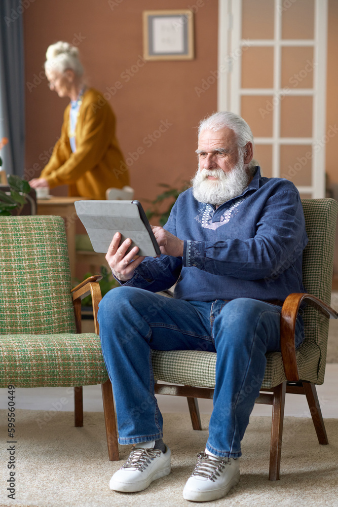 Vertical image of senior bearded man using tablet pc while sitting on armchair in the living room at home with his wife in background