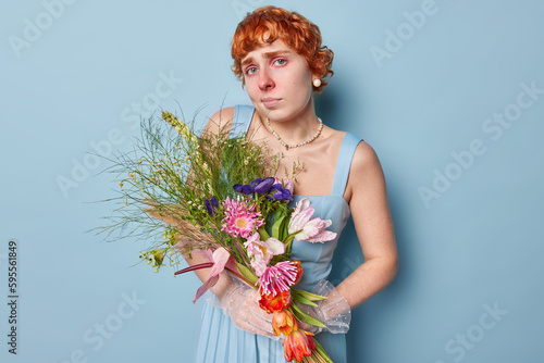 Upset displeased redhead woman in dress holds big bouquet of wild flowers has red itchy eyes and nose suffers from allergy poses against blue background. Health problems and allergic reaction concept