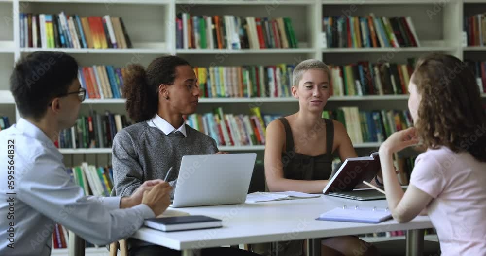 Four young attractive multi ethnic students sitting at table with workbooks and laptop in library, preparing common project, making assignment, share ideas and opinions engaged in team-work. Education Four young attractive multi ethnic students sitting at table with workbooks and laptop in library, preparing common project, making assignment, share ideas and opinions engaged in team-work. Education