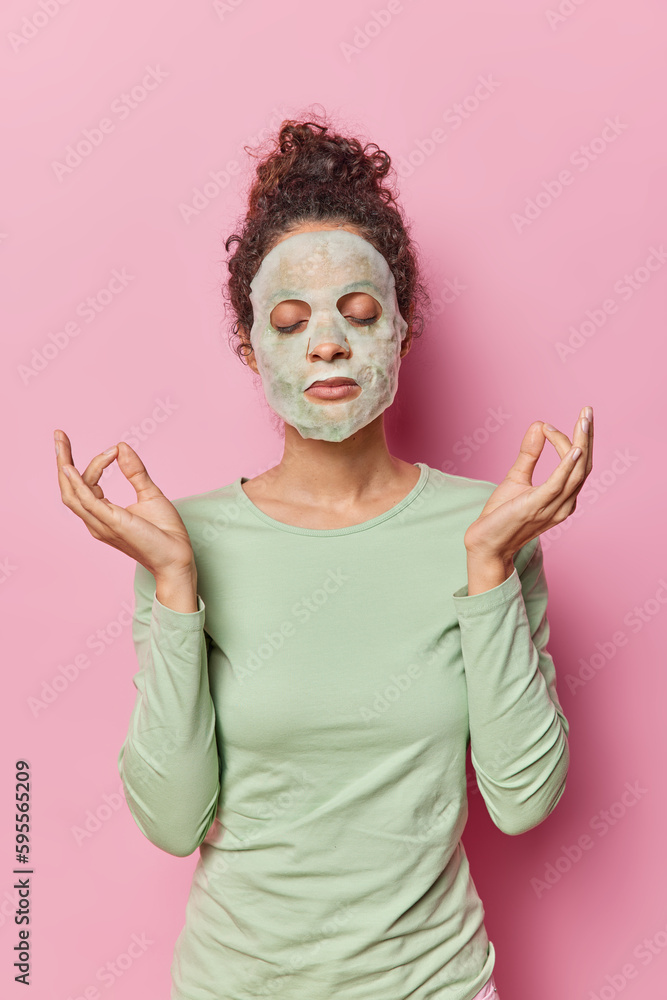 Relaxed dark haired woman applies moisturising soap mask on face keeps eyes closed meditates to relax undergoes beauty procedures wears casual jumper isolated over pink background. Beauty concept