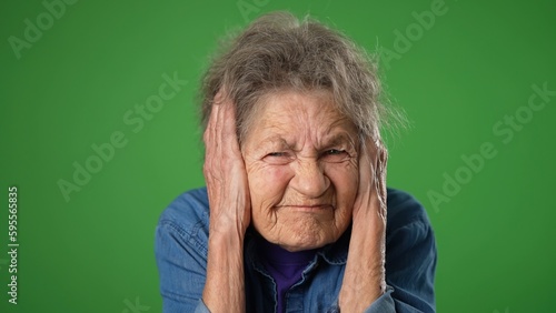 Unhappy irritated frustrated sad angry toothless old elderly senior woman with wrinkled skin, grey hair closed eyes cover ears do not want to listen scream on green screen background studio portrait