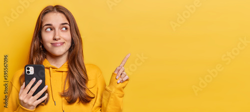 Horizontal shot of good looking teenage girl points fore finger at upper left corner has news about bargains holds modern smartphone in hand wears sweatshirt isolated over vivid yellow background