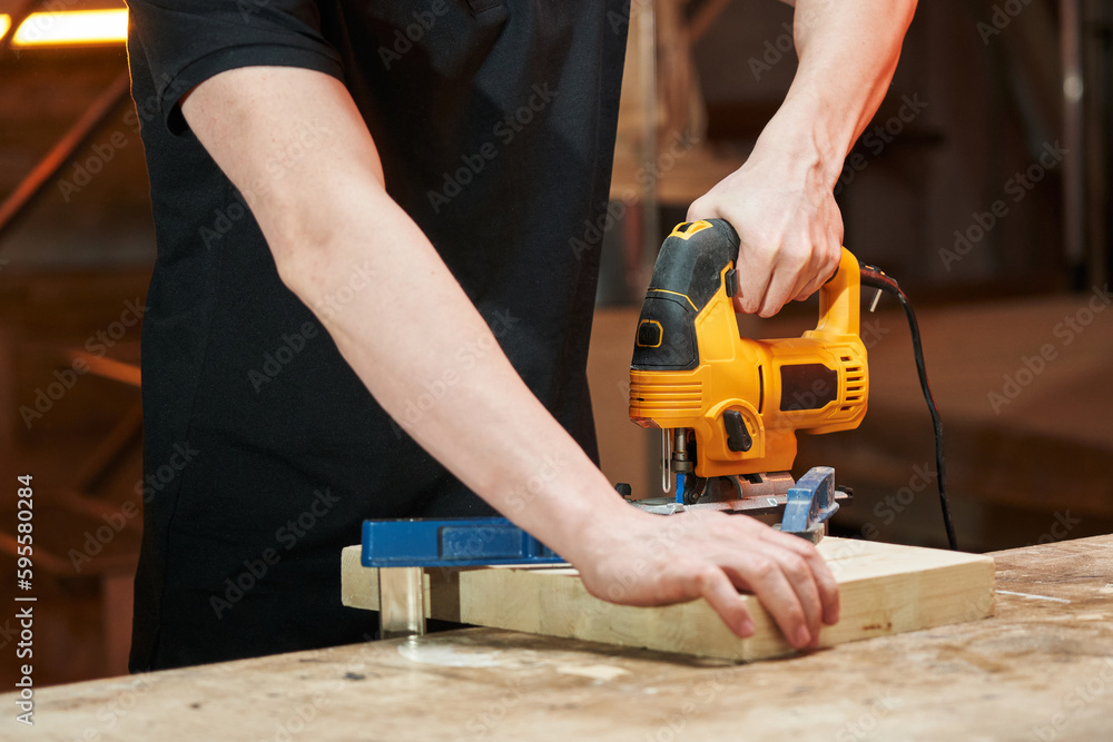 caucasian worker saws a wooden blank beam fixed with clamps to a workbench with an electric jigsaw caucasian worker saws a wooden blank beam fixed with clamps to a workbench with an electric jigsaw