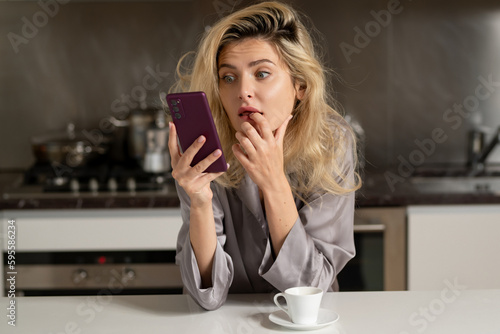 Surprised woman sitting in the kitchen watching phone in the morning and chatting smartphone.