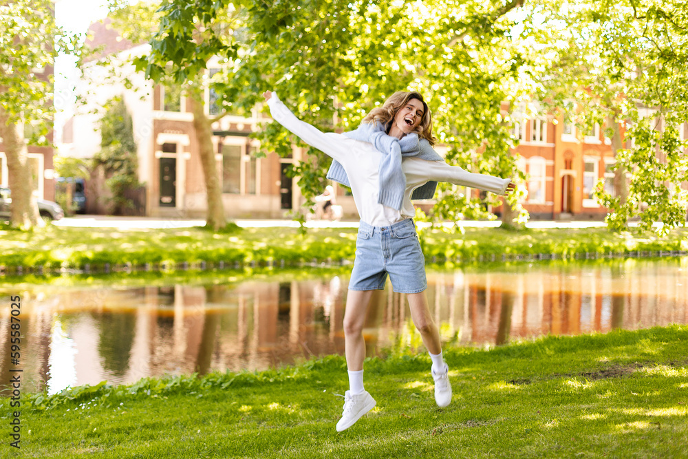 Portrait of beautiful woman smiling and jumping in park at sunny day. Outdoor portrait of a smiling curly blonde girl. Happy cheerful girl laughing at park wear sweater, white long sleeves, shorts