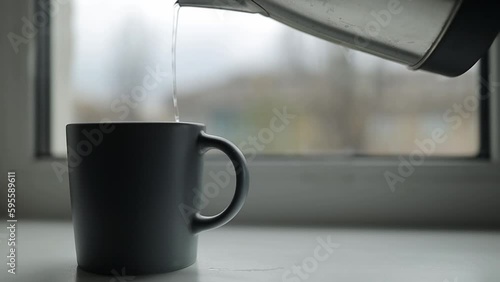 pours boiling water from an electric kettle into coffee in a gray mug. pour hot boiled water from an electric kettle into a mug. The kettle pours boiling water into a cup on the windowsill.