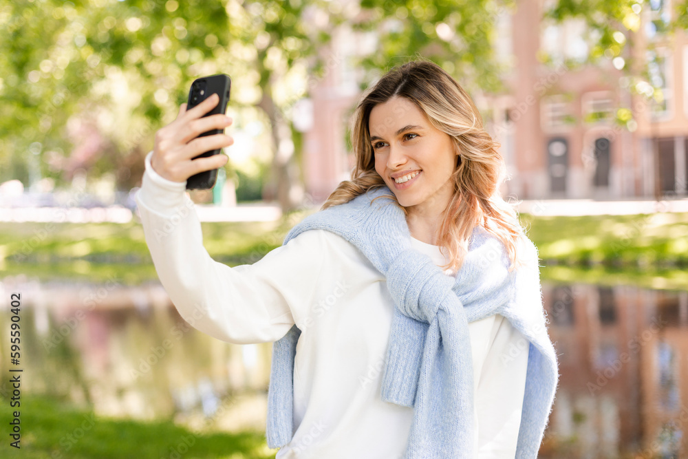 Image of smiling young curly hair blonde woman taking selfie photo holding her phone while walking in park near river. Girl make video call using her phone, look happy and smiling. Image of smiling young curly hair blonde woman taking selfie photo holding her phone while walking in park near river. Girl make video call using her phone, look happy and smiling.