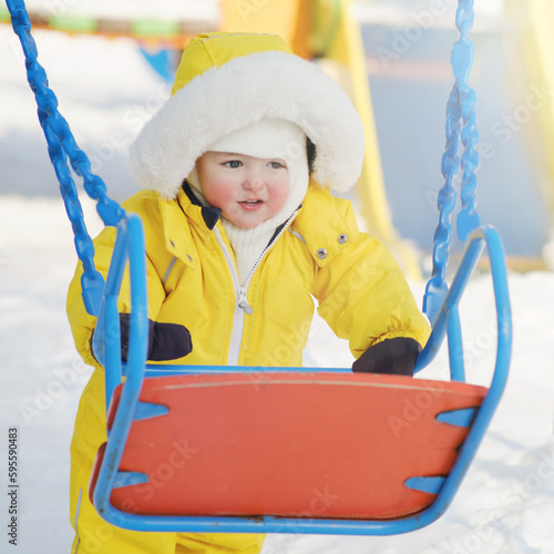 Happy toddler baby himself climbs on the swing in the winter playground. Baby boy in yellow snowsuit swinging in kindergarten. Kid age one year eight months