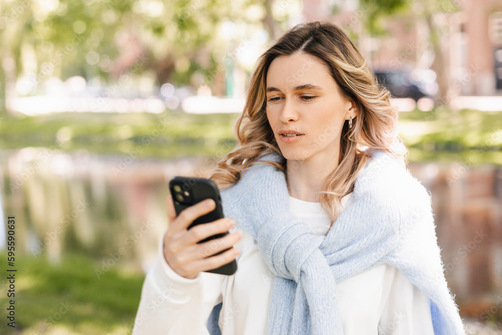 Young stylish curly blonde woman writes message on smartphone walking on park near river, hipster tourist traveling in european town and planning trip using mobile phone. Girl wear sweater and t-shirt Young stylish curly blonde woman writes message on smartphone walking on park near river, hipster tourist traveling in european town and planning trip using mobile phone. Girl wear sweater and t-shirt