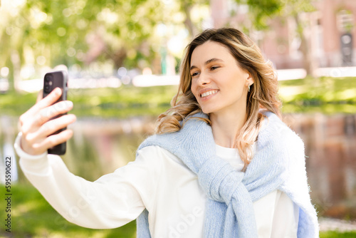 Image of smiling young curly hair blonde woman taking selfie photo holding her phone while walking in park near river. Girl make video call using her phone, look happy and smiling.