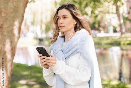 Young stylish curly blonde woman writes message on smartphone walking on park near river, hipster tourist traveling in european town and planning trip using mobile phone. Girl wear sweater and t-shirt
