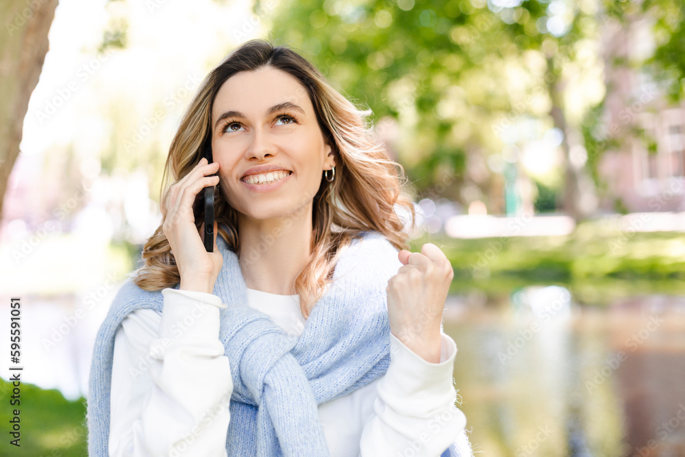 Young happy blonde woman with curly hair received good unexpected news on the phone call in the park. Work promotion, Job offer, great news, girl make win gesture wear blue sweater, white t-shirt.