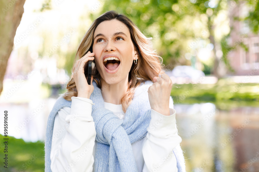 Young happy blonde woman with curly hair received good unexpected news on the phone call in the park. Work promotion, Job offer, great news, girl make win gesture wear blue sweater, white t-shirt. Young happy blonde woman with curly hair received good unexpected news on the phone call in the park. Work promotion, Job offer, great news, girl make win gesture wear blue sweater, white t-shirt.