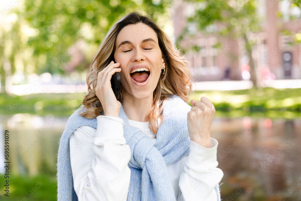 Young happy blonde woman with curly hair received good unexpected news on the phone call in the park. Work promotion, Job offer, great news, girl make win gesture wear blue sweater, white t-shirt.