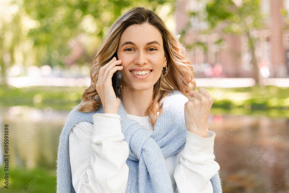 Young happy blonde woman with curly hair received good unexpected news on the phone call in the park. Work promotion, Job offer, great news, girl make win gesture wear blue sweater, white t-shirt. Young happy blonde woman with curly hair received good unexpected news on the phone call in the park. Work promotion, Job offer, great news, girl make win gesture wear blue sweater, white t-shirt.