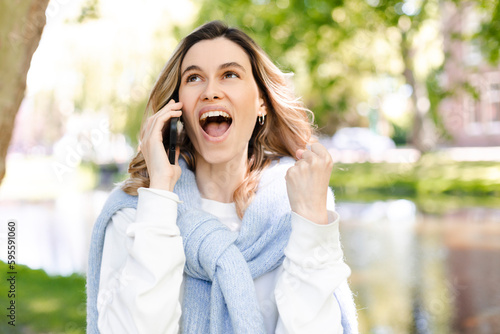 Young happy blonde woman with curly hair received good unexpected news on the phone call in the park. Work promotion, Job offer, great news, girl make win gesture wear blue sweater, white t-shirt.