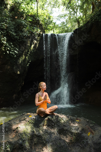 The girl is meditating, her hands are folded in namaste, in the jungle against the backdrop of a waterfall, dressed in a yellow swimsuit.