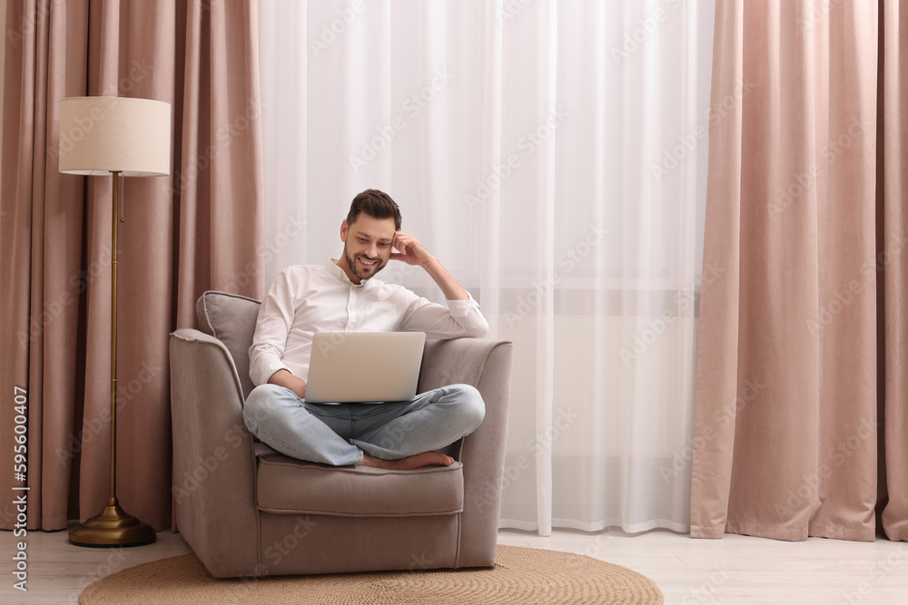 Smiling man holding laptop on armchair near window with beautiful curtains at home. Space for text