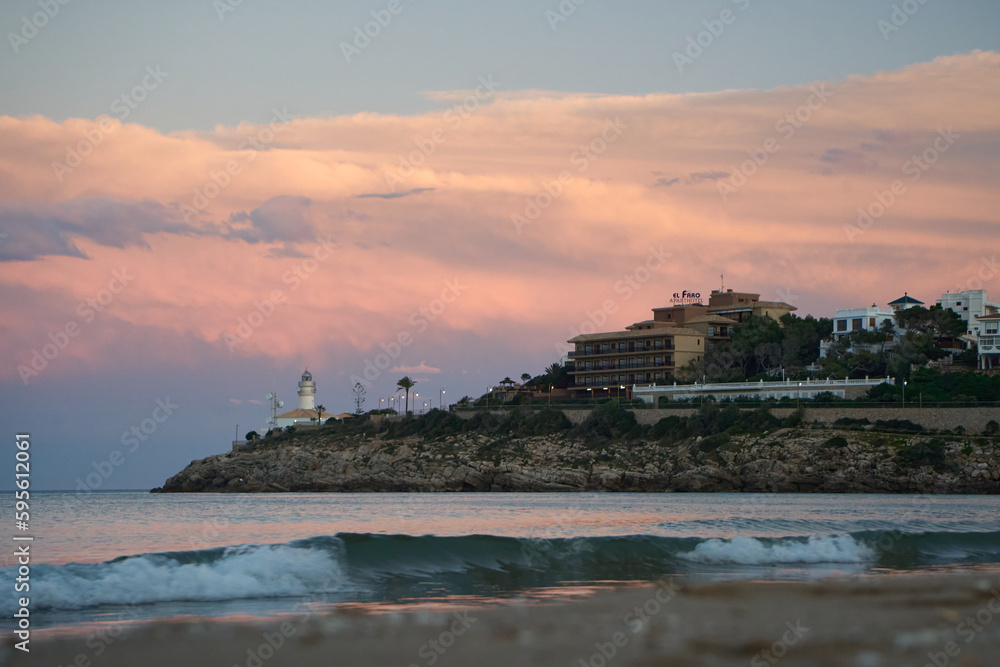 a lighthouse on the beach at dusk with a pink and white cloud in the sky
