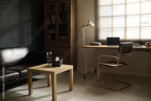 Workplace of psychologist by window and small square wooden table by black comfortable leather couch standing by wall in psychotherapy office © pressmaster