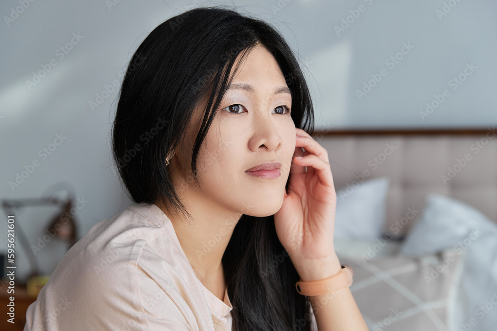 Close up serious asian woman in casual clothes looking out the window sitting on the bed at home Close up serious asian woman in casual clothes looking out the window sitting on the bed at home