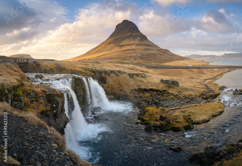 This panoramic shot captures the beauty of Kirkjufellsfoss waterfall and Kirkjufell mountain in Iceland. The rugged landscape and changing weather conditions during sunset