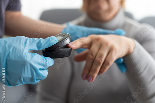 Medical help at home. Woman doctor in a medical mask measures the patient's pulse and oxygen saturation to middle aged woman using a pulse oximeter while sitting on sofa, healthcare concept.