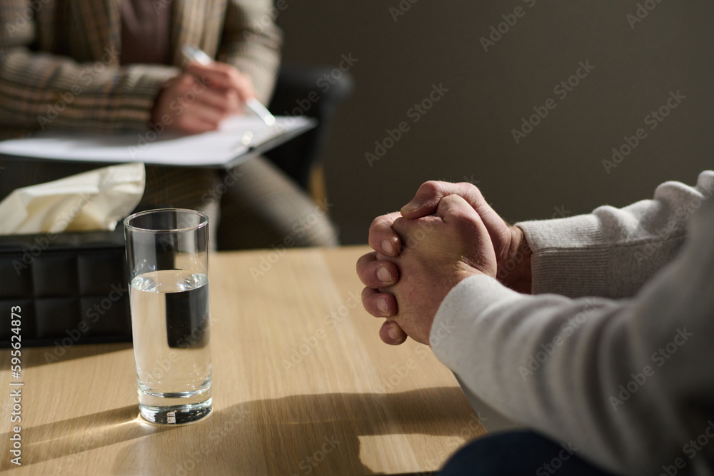 Clasped hands of stressed mature man sitting by table with glass of water and box with tissues in front of psychologist making notes