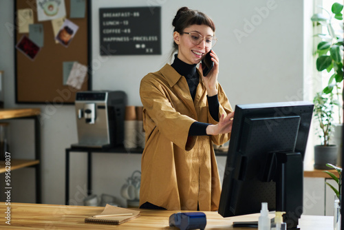 Print op canvas Young smiling female clerk talking with client on smartphone and entering order