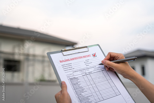 An engineer hand is using a pen checking on the building inspection report form to QC building quality of the house (background). Export industrial working scene concept. Close-up and selective focus.