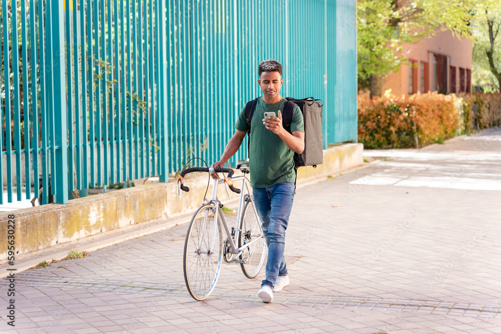 Fototapeta premium Cheerful ethnic man with bicycle walking with backpack delivering food