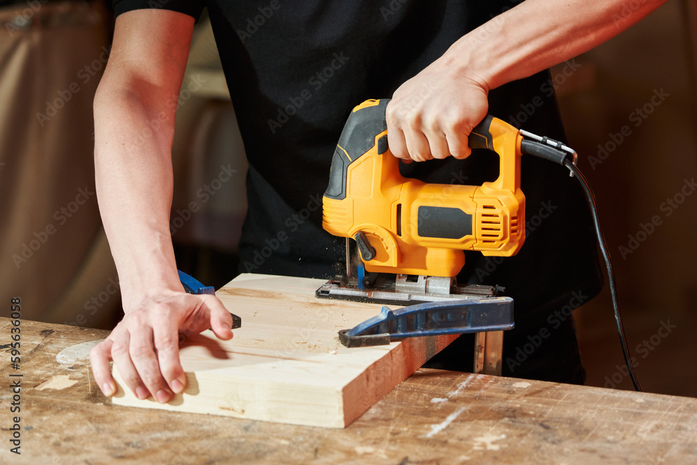 the hands of a Caucasian worker using an electric jigsaw saw off a part of a wooden board fixed with clamps to a workbench the hands of a Caucasian worker using an electric jigsaw saw off a part of a wooden board fixed with clamps to a workbench