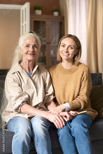 Vertical image of happy senior mom sitting on sofa with her adult daughter and they smiling at camera together