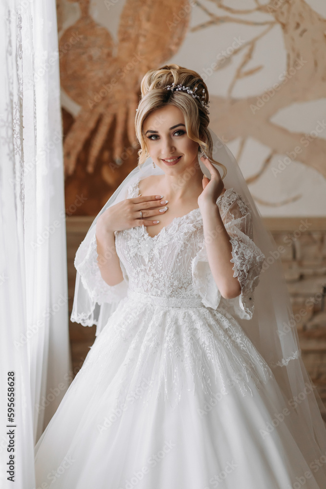 Fototapeta premium A blonde bride in a white dress and a long veil is posing by the window. Beautiful hands. French manicure. Beautiful hair and make-up. Diadem. Daylight