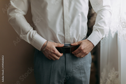 Details. A man in a white shirt and gray pants adjusts his stylish belt. Masculine style. Fashion. Business