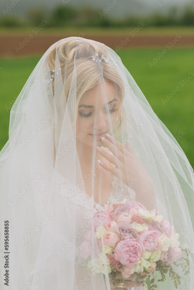Portrait of the bride. The bride is standing with a bouquet in a white dress covered with a veil, in a green field. A wonderful dress. Pretty Woman. Wedding photo