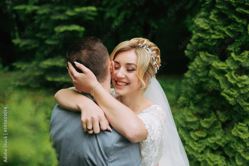 Portrait of the bride and groom standing on the background of green trees, embracing. The bride smiles sincerely. Stylish groom. Fashion and style. Beautiful bride